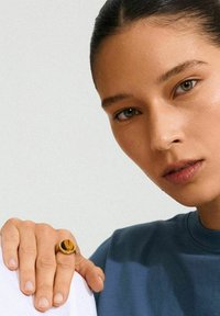 Zlatni prsten s velikim ovalnim smeškim tigrovim okom. Model nosi plavu majicu, a blaga pozadina ističe detalje prstena.