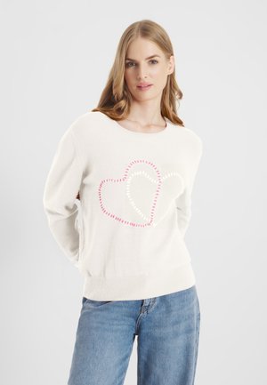 Młoda kobieta z długimi blond włosami, ubrana w biały sweter z dwoma obszytymi różowymi i białymi konturami serc, do tego niebieskie dżinsy.