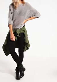 Pull en tricot gris avec manches chauve-souris, leggings noirs et bottines. Chemise verte nouée à la taille. Textures lisses et couleurs contrastées.