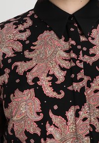 Svart skjorta med paisleymönster i rosa och beige. Strukturerad krage och knappar vid halsen. Slät tygtextur.