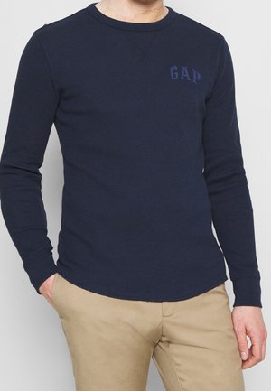 Mężczyzna ubrany w granatową koszulę z długim rękawem z logo "GAP" na piersi i spodnie koloru khaki, stojący z jedną ręką w kieszeni na jasnym tle.