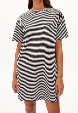 Femme portant une robe à manches courtes à rayures horizontales noires et blanches avec une petite poche poitrine, debout devant un fond clair uni.
