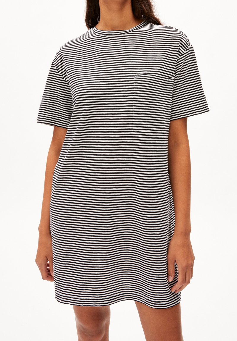 Femme portant une robe à manches courtes à rayures horizontales noires et blanches avec une petite poche poitrine, debout devant un fond clair uni.