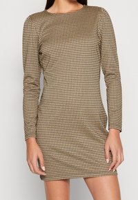 Robe mini à manches longues en tissu texturé avec un motif à carreaux beige et noir, présentant un col rond et une silhouette ajustée.