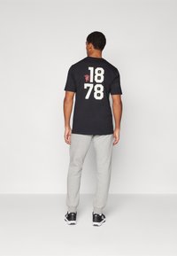 Črna bombažna majica s velikimi belimi številkami "1878" in "MUFC" natisnjenimi na hrbtni strani, v kombinaciji s sivo trenirka in črnimi supergami.
