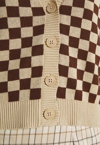 Braun-beige karierte Strickjacke mit geripptem Saum, ausgestattet mit vier großen, hellen Knöpfen. Weiche Stricktextur.