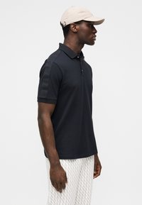 Tumesinine polopluus kahe nupuga kraega, lühikeste varrukatega ja toonitoonis logoga varrukatel. Sobitatud helekirjuliste altosade ja beežiga torknimutsuga.