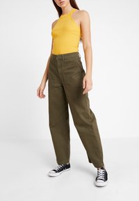 Top halter giallo a coste abbinato a pantaloni a vita alta verde oliva. Sneakers in tela nere completano l'outfit. Design semplice e vestibilità casual.