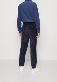 Navy corduroy trousers with a back pocket, paired with a blue button-up shirt. Model wears white sneakers, presenting a casual look.