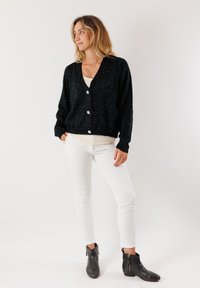 Cardigan nero in maglia con texture scintillante e bottoni frontali, indossato sopra una maglietta chiara e abbinato a jeans bianchi e stivaletti neri.