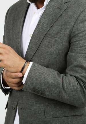 Blazer grigio a scacchi con un design strutturato, caratterizzato da un colletto a scollo e polsini con tre bottoni, indossato sopra una camicia bianca. Braccialetto nero visibile.