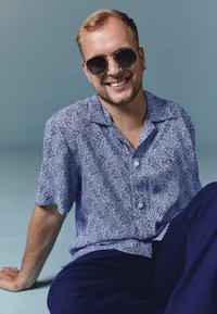 Chemise à manches courtes bleu avec des motifs tourbillonnants blancs, associée à un pantalon bleu foncé. Accessoirisée avec des lunettes de soleil rondes.