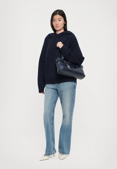 Navy hoodie with oversized fit, light blue flared jeans, and white pointed heels. Dark blue handbag with strap and key detail.