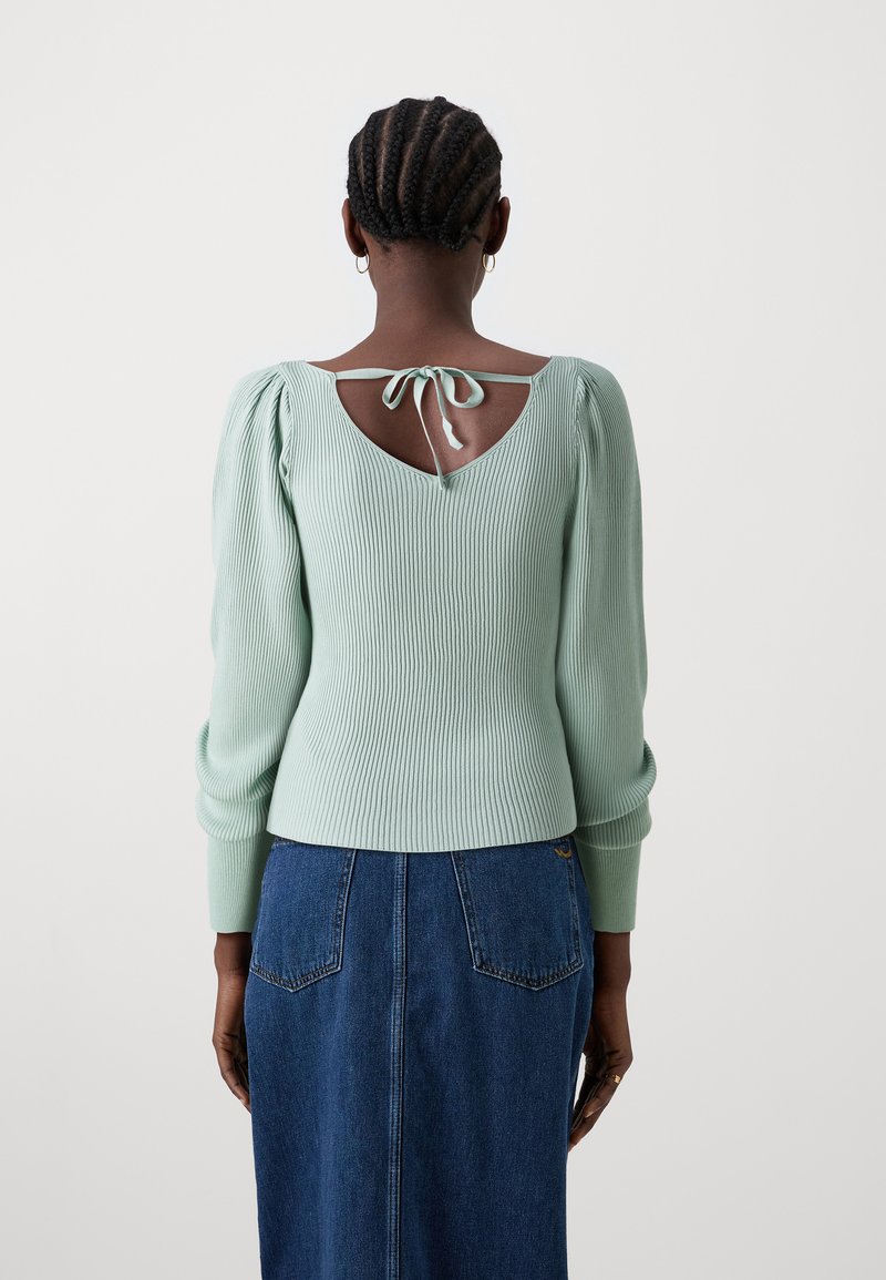 Maglione a coste verde menta con maniche a sbuffo, schiena scoperta con chiusura a lacci, abbinato a una gonna di jeans blu e accessori minimi.