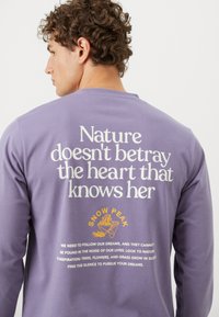 Purple long-sleeve shirt featuring white and yellow text on the back. Material appears soft, with a relaxed fit and crew neckline.