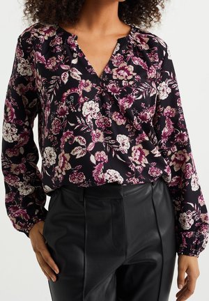 Kvindelig iført en sort bluse med lilla og hvide blomsterprint og lange ærmer, parret med sorte læderbukser.