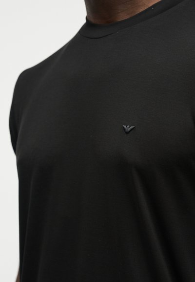 Κοντινό πλάνο ενός ατόμου που φοράει μια απλή μαύρη μπλούζα με ένα μικρό, μαύρο κεντητό λογότυπο στο αριστερό μέρος του στήθους.