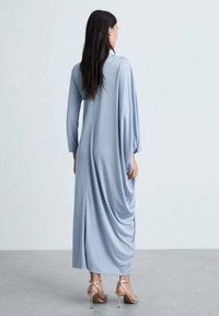 Long, light blue dress with a draped design, long sleeves, and side slits. Smooth fabric texture paired with strappy gold heels.