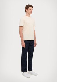 T-shirt in cotone beige chiaro, maniche corte, collo rotondo. Abbinato a jeans in denim blu scuro e sneaker bianche, design minimale senza decorazioni visibili.