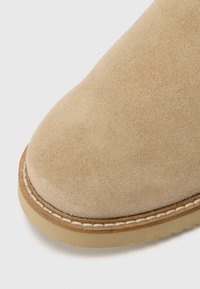 Botas de ante beige en primer plano, con punta redondeada, detalles de costura y suela de goma marrón.