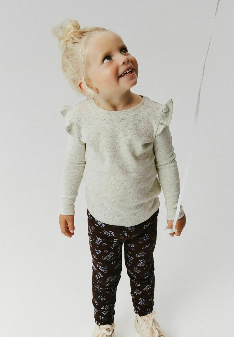 Jeune fille blonde avec un chignon tenant une ficelle de ballon, portant un pull blanc à volants et un pantalon à fleurs foncé, regardant vers le haut et souriant.