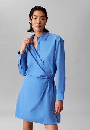 Junge Frau mit kurzen schwarzen Haaren trägt ein langärmliges, hellblaues Wickelkleid mit Kragen und steht vor einem schlichten grauen Hintergrund.