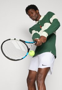 Zielona i kremowa w paski koszulka polo, białe szorty z logo Nike. Gracz trzyma rakietę tenisową i przygotowuje się do uderzenia w żółtą piłkę tenisową.