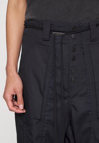 Schwarze Hose aus strukturiertem Stoff, ausgestattet mit großen Taschen, einem Kordelzugbund und metallischen Druckknöpfen entlang der Vorderseite.