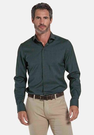 Man met donker haar en baard, draagt een donkergroen overhemd met knopen en beige broek met een donkere riem, staand tegen een effen achtergrond.