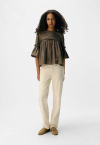 Kvinde med krøllet hår iført en løs olivenfarvet bluse med flæser, off-white bukser og lysebrune loafers, stående foran en hvid baggrund.