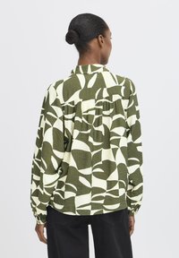 Kvinde med mørkt hår i en knold iført en langærmet bluse med grønne og hvide abstrakte mønstre og sorte bukser, som vender væk.