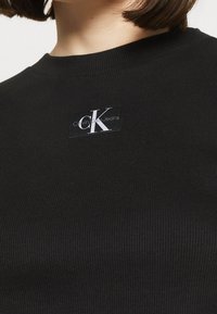 Černý žebrovaný svetr s plochou nášivkou loga Calvin Klein Jeans, která je výrazně umístěna. Texturovaná látka, kulatý výstřih.