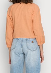 Blusa arancione cropped con maniche a sbuffo, texture non decorata. Abbinata a jeans azzurri a vita alta con una piccola toppa a triangolo.