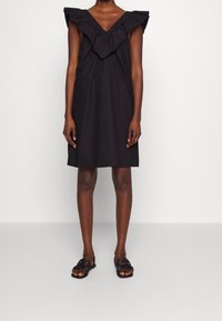 Schwarzes Kleid mit rüschiertem V-Ausschnitt und kurzen Ärmeln. Leichtes Material, knielanges Design. Model kombiniert es mit schwarzen Sandalen mit Riemchen.