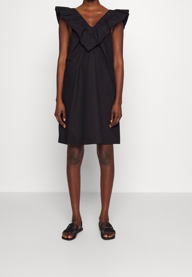 Schwarzes Kleid mit rüschiertem V-Ausschnitt und kurzen Ärmeln. Leichtes Material, knielanges Design. Model kombiniert es mit schwarzen Sandalen mit Riemchen.