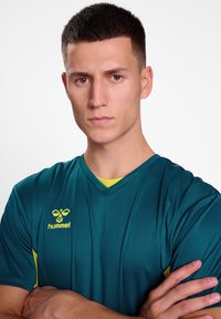 Teal sportströja med korta ärmar, V-ringad design och gula detaljer. Har "hummel"-logotypen på vänster bröst. Slät tygtextur.