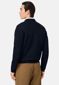 Tmavomodrý pulóver s golierom, so vzorovanými manžetami a pásom, zobrazený zozadu, v kombinácii s pieskovými nohavicami. Hladká textúra.