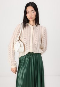 Blusa de encaje de manga larga en crema con cierre de botones, combinada con una falda de satén verde esmeralda con pliegues. Accesorio con un bolso blanco.