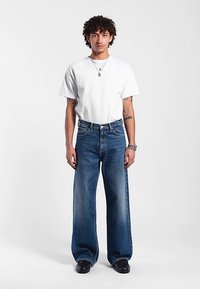 Weißes T-Shirt mit kurzen Ärmeln, kombiniert mit weit geschnittenen blauen Denim-Jeans. Das Outfit besticht durch eine lockere Passform und ein minimalistisches Design. Glatte Textur.