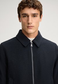 Marineblauwe wollen mix jas met kraag, ritssluiting en zijzakken. Getextureerde stof met een getailleerde pasvorm.