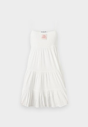 Femme portant une robe blanche sans manches avec un corsage smocké, des chaussettes blanches, des bottes rouges à lacets et des lunettes de soleil foncées sur un fond uni.