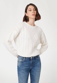 Witte gebreide trui met een textuur van bladeren, ronde halslijn en lange mouwen, gecombineerd met blauwe denim jeans.