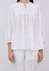 Top in cotone bianco con scollatura rotonda, dettaglio a chiave, polsini elasticizzati e motivi geometrici a occhiello. Abbinato a pantaloni bianchi a occhiello.