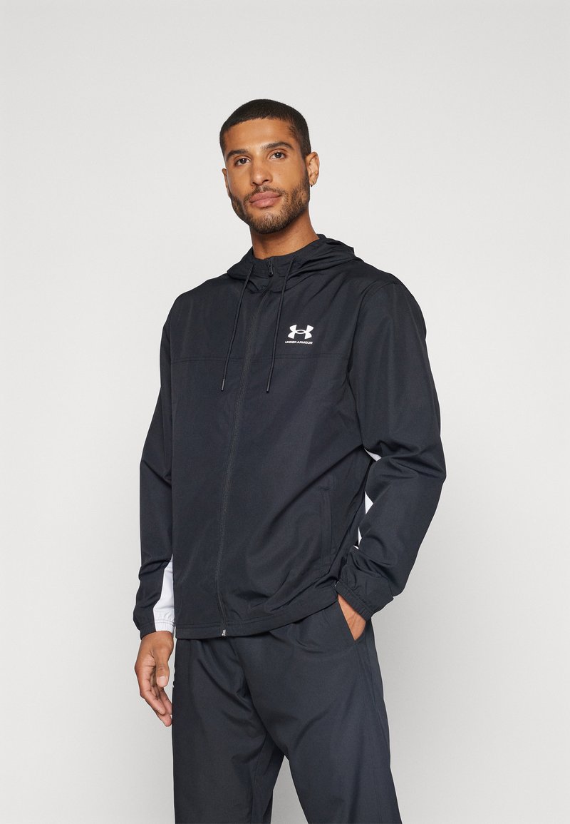 Veste noire zippée avec capuche, logo blanc sur la poitrine et accents blancs contrastants sur les manches. Tissu lisse avec un léger brillant.