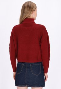 Roter gestrickter Pullover mit hohem Kragen und Kabelstrickmuster an den Ärmeln, kombiniert mit einem dunklen Jeans-Minikleid mit hinteren Taschen.