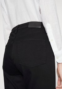 Svarta jeans med en läderlogotyp på midjan. Materialet är mjukt och stretchigt, med två bakfickor och subtila sömdetaljer.