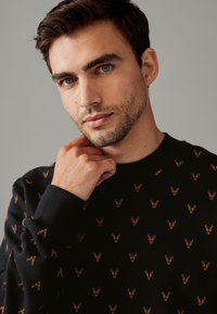 Maglione nero con un motivo ripetitivo di piccoli teschi di cervo marroni. Ha un collo rotondo e polsini e orlo ribattuti. Morbido al tatto.