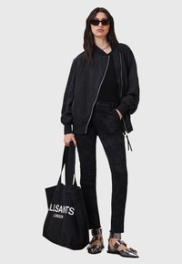 Chaqueta bomber negra sobre una camiseta negra, combinada con pantalones de camuflaje negros. Los accesorios incluyen un bolso tote negro y zapatos de tobillo metálicos.