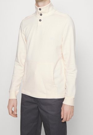 Man draagt een crèmekleurige pullover met lange mouwen, een hoge, dichtgeknopte kraag en een voorvak, gecombineerd met een donkere broek.