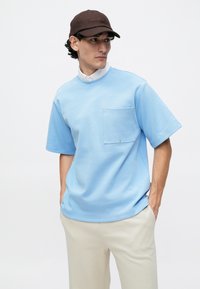 T-shirt oversized azul claro com mangas curtas, detalhe de bolso e decote redondo. Usado com calças bege e um boné castanho.